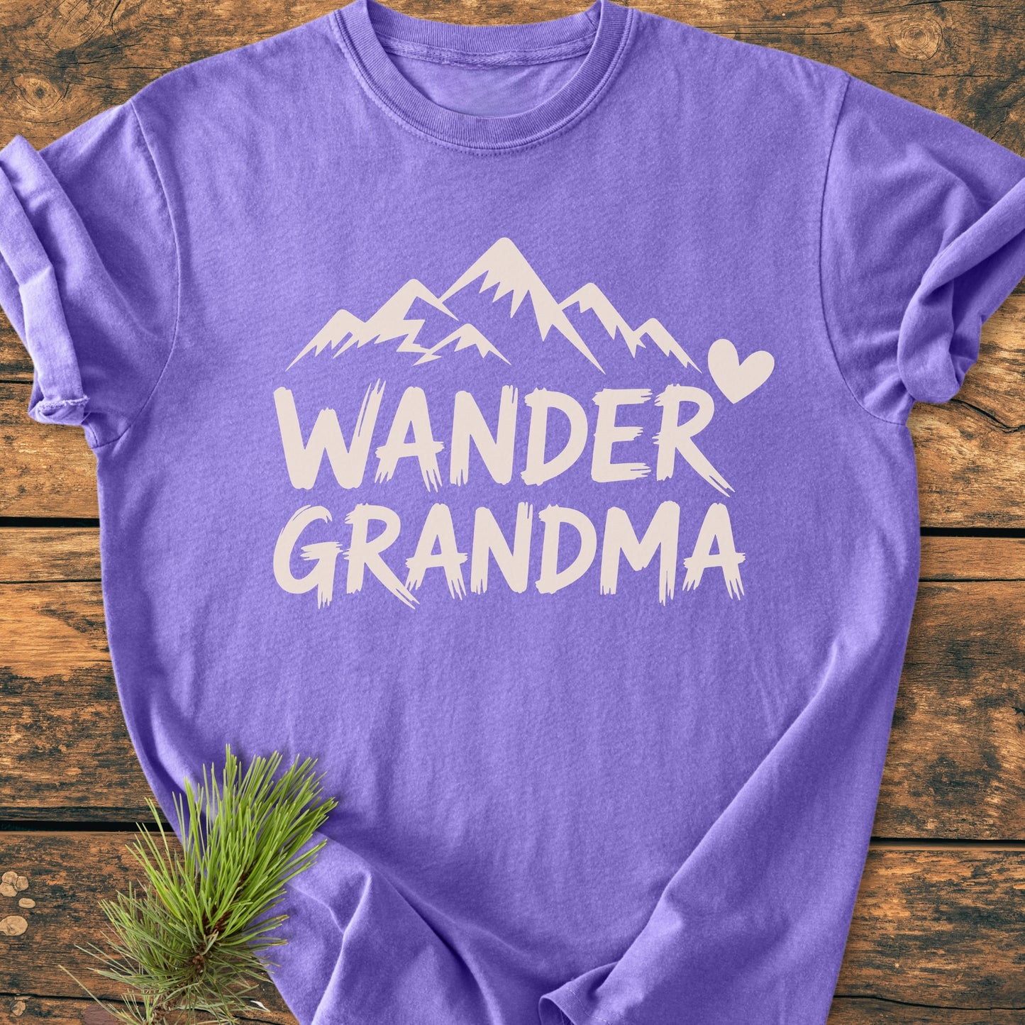 Wander Grandma - Vintage Heavyweight Tee featuring a stylized mountain and heart design. Laid flat on wood, showcasing its rugged charm and durable, relaxed fit. Includes pine sprig accent.