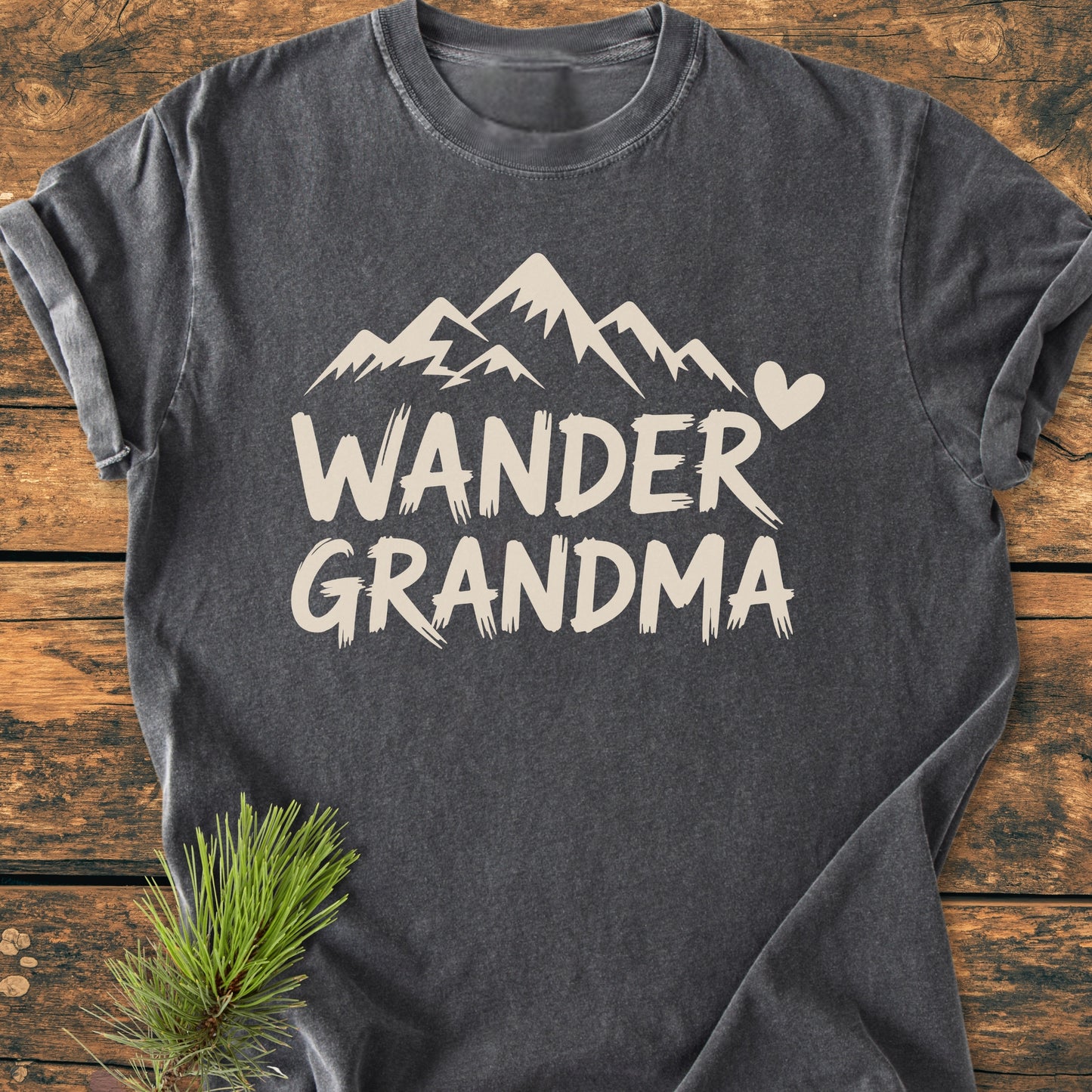 Wander Grandma - Vintage Heavyweight Tee featuring a stylized mountain range and heart printed on a dark gray short-sleeved shirt, displayed on a rustic wood surface with a pine sprig beside it.