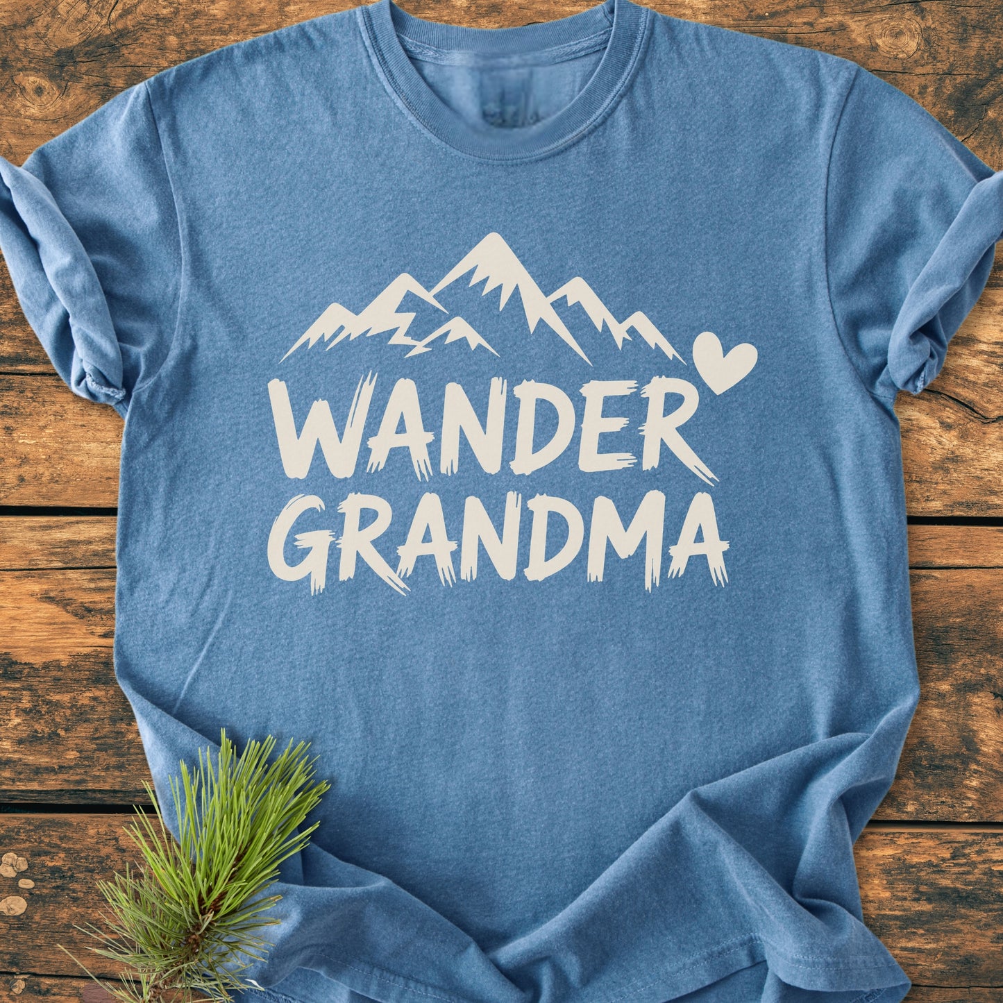 Heather-blue Wander Grandma - Vintage Heavyweight Tee featuring stylized mountain peaks and a heart graphic, laid on wood with a pine sprig, highlighting its soft, durable, relaxed-fit design.