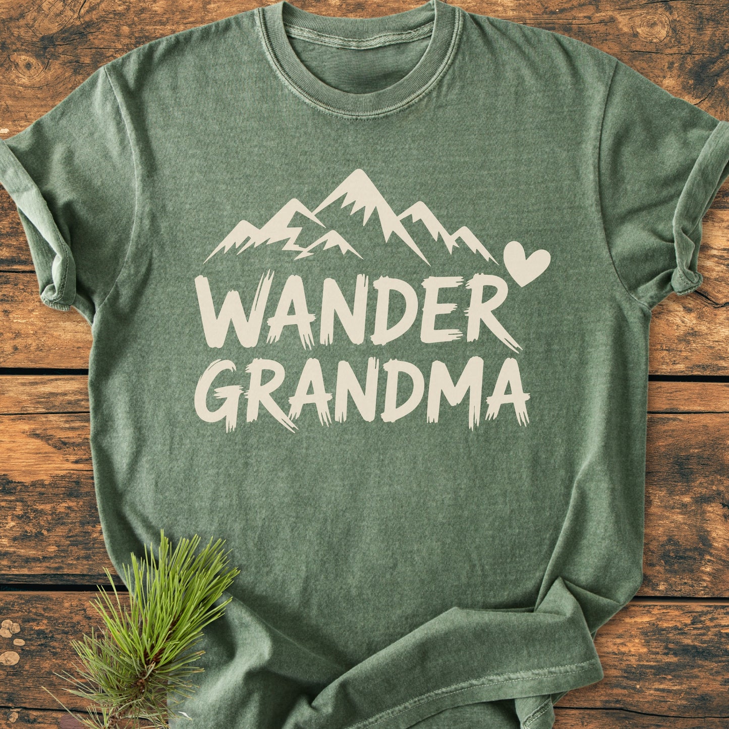 Wander Grandma - Vintage Heavyweight Tee featuring a stylized mountain and heart design in off-white on a green heathered fabric, laid flat with a pine branch accent for an outdoorsy touch.
