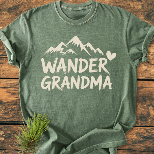 Wander Grandma - Vintage Heavyweight Tee featuring a stylized mountain and heart design in off-white on a green heathered fabric, laid flat with a pine branch accent for an outdoorsy touch.