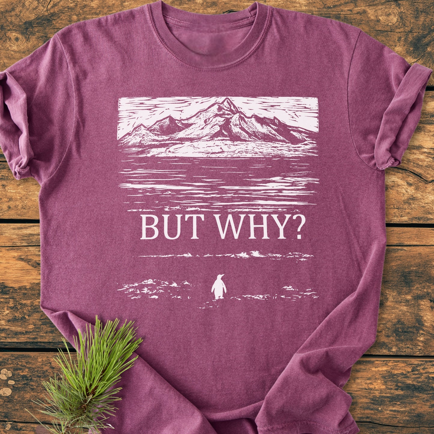 The But Why - Vintage Heavyweight Tee features a white stylized mountain landscape and penguin graphic on a purple-mauve T-shirt, laid flat on a wooden surface with a pine sprig beside it.