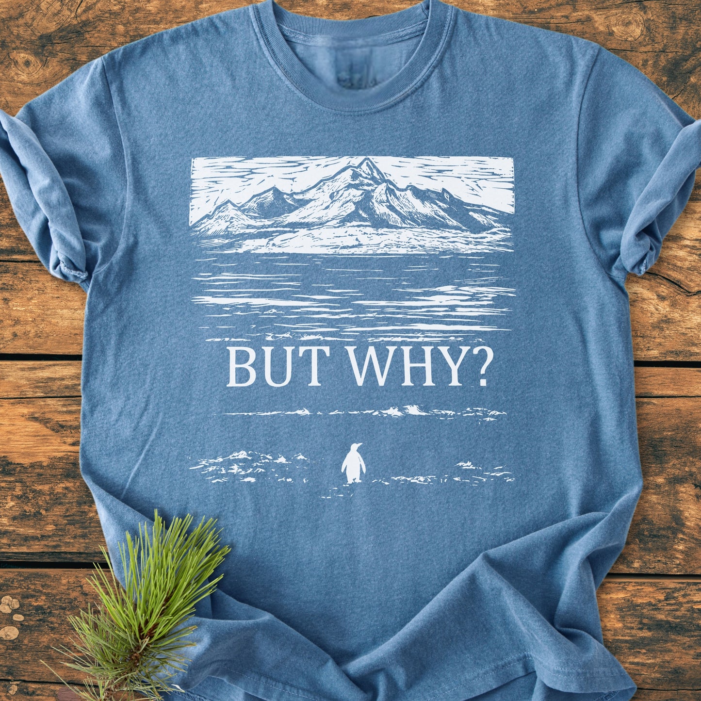 But Why - Vintage Heavyweight Tee features a heather-blue shade with a woodcut-style mountain scene and BUT WHY? text, accented by a penguin silhouette. Styled with a pine sprig.