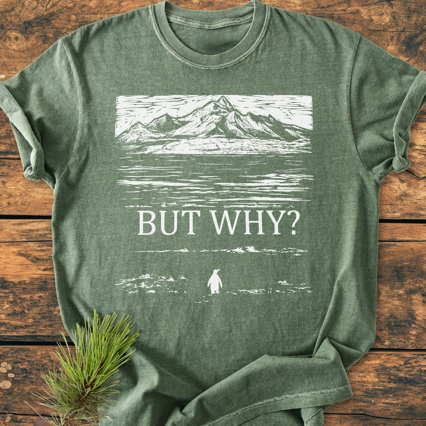 But Why - Vintage Heavyweight Tee featuring a linocut-style mountain and penguin design, laid flat with a pine sprig on a wooden surface, showcases a relaxed fit and durable stitching.