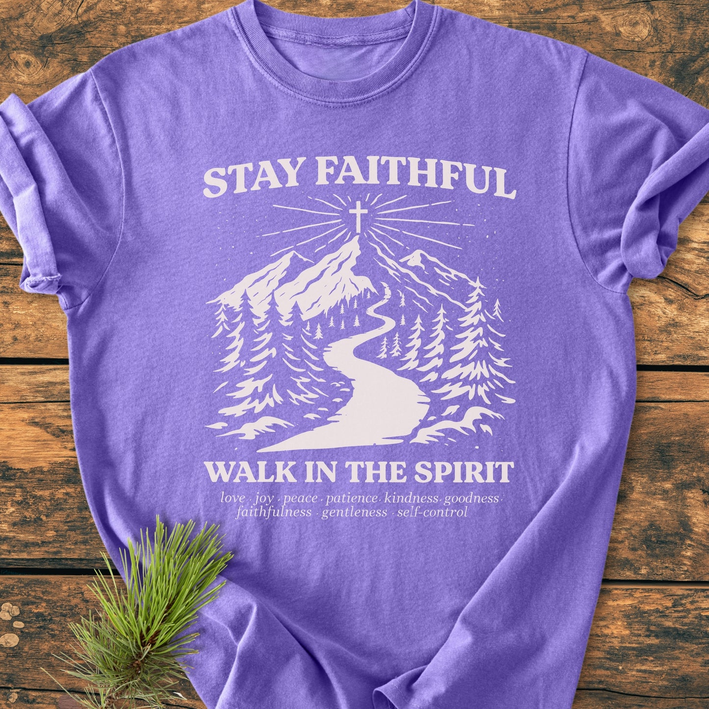 Stay Faithful - Vintage Heavyweight Tee features white screen-printed text and mountain scene on a purple T-shirt, laid on a wooden surface with a pine sprig, emphasizing spiritual themes and durability.