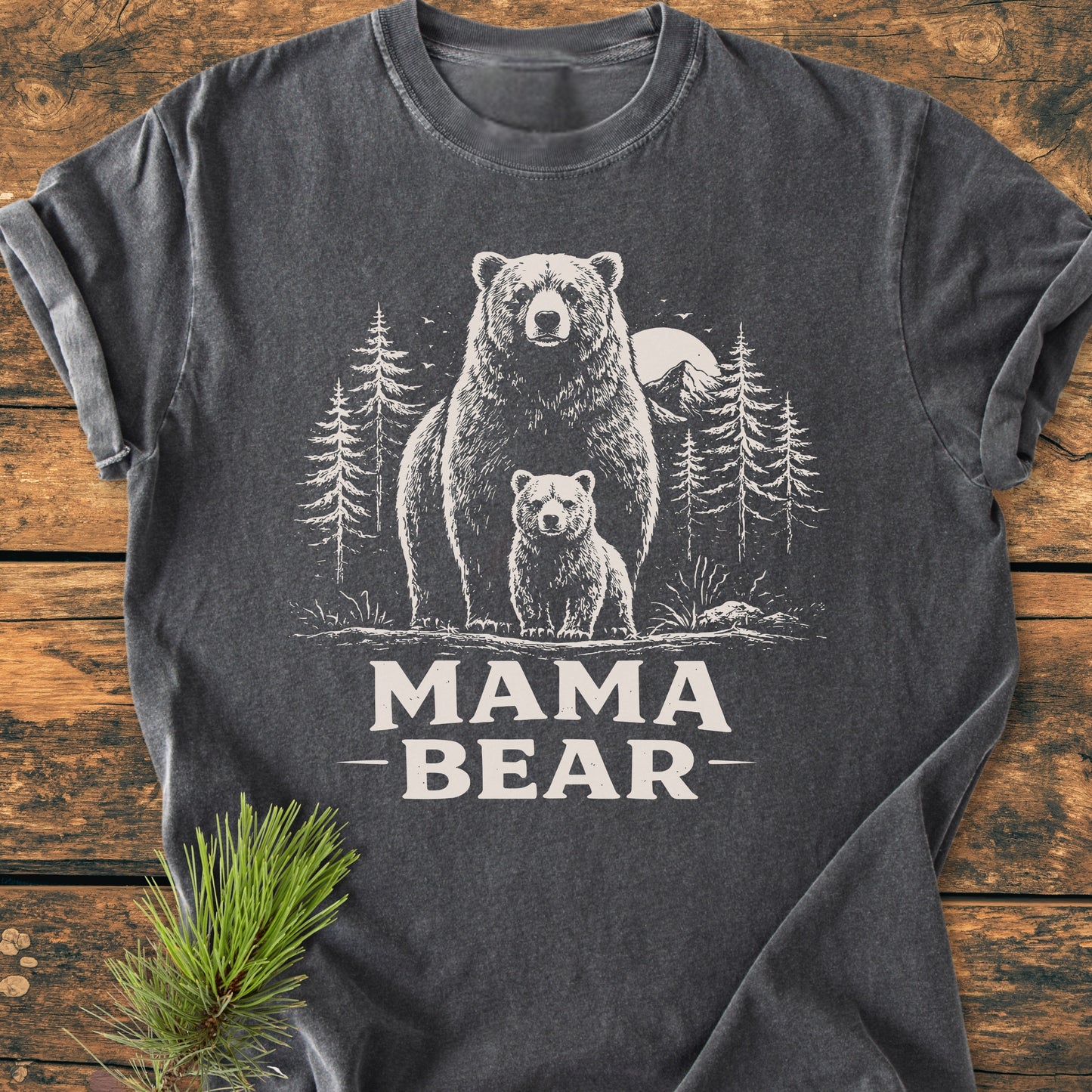 Mama Bear - Vintage Heavyweight Tee featuring a hand-drawn bear scene with MAMA BEAR text on a dark-gray shirt, laid on wood, with a pine sprig accentuating the design.