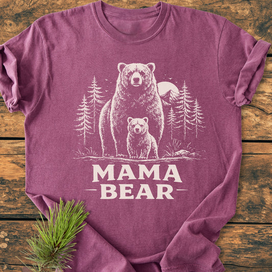 Mama Bear - Vintage Heavyweight Tee featuring a white bear and cub design amidst trees and sun, folded on a wooden surface with pine needles beside, highlighting its soft, durable 100% ring-spun cotton.
