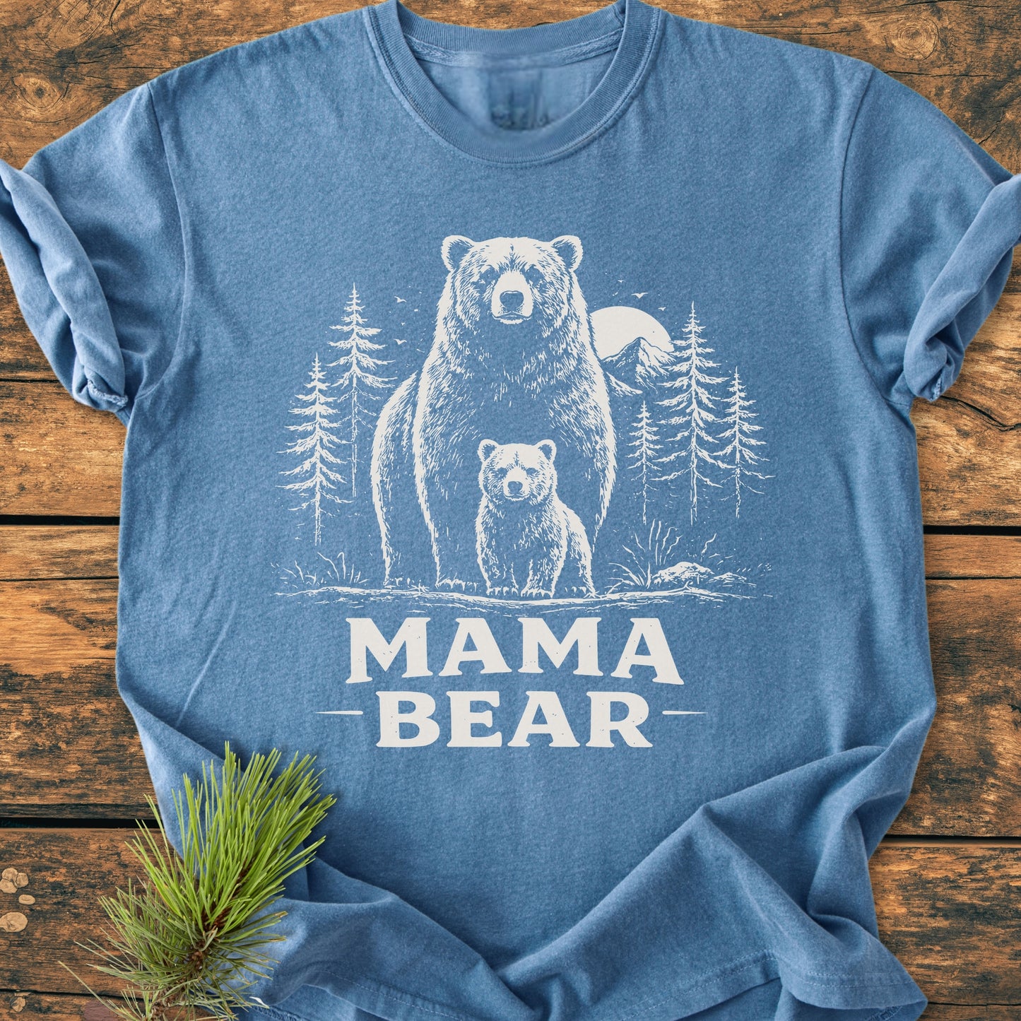 Mama Bear - Vintage Heavyweight Tee features a hand-drawn bear and cub illustration in a forest with a sun. The blue tee is folded on wood, accented by a pine sprig.