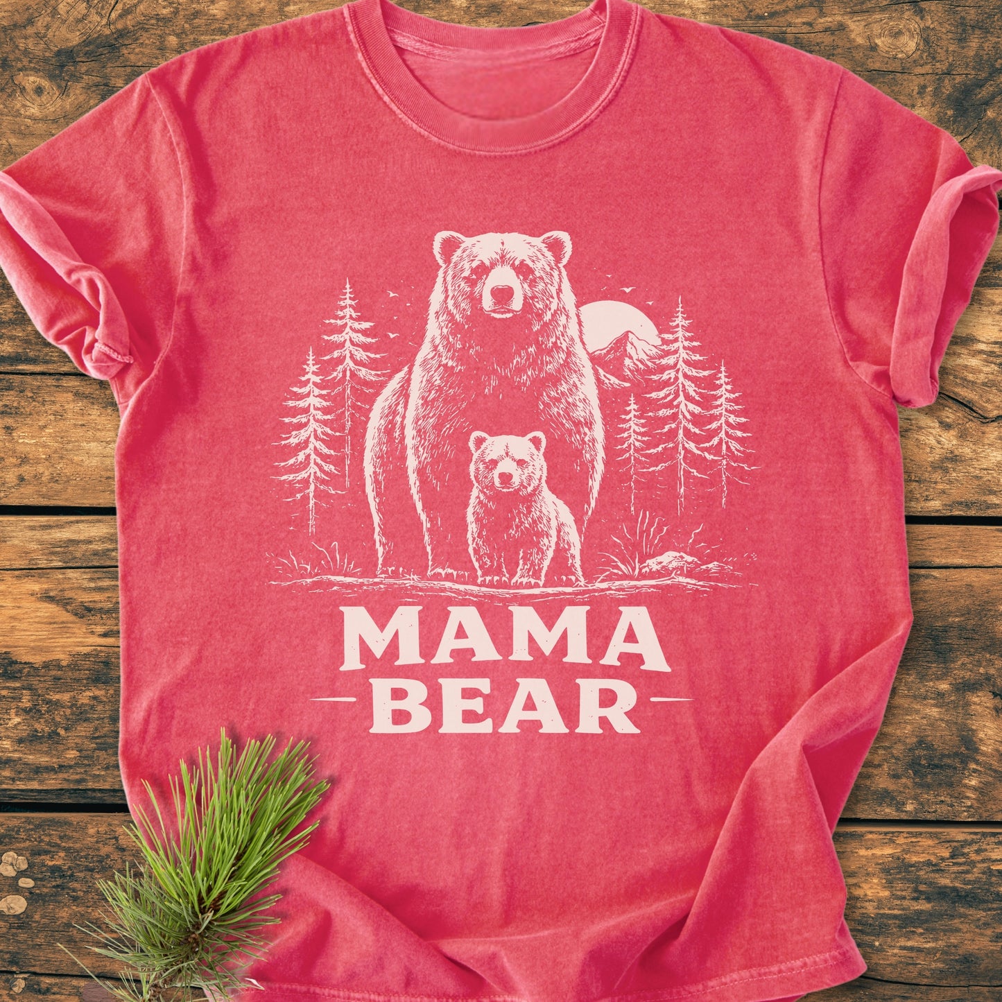Mama Bear - Vintage Heavyweight Tee features a bear illustration with cub and pine trees against a sun backdrop, laid on a wooden surface. Styled with pine needles, highlighting its relaxed, durable design.
