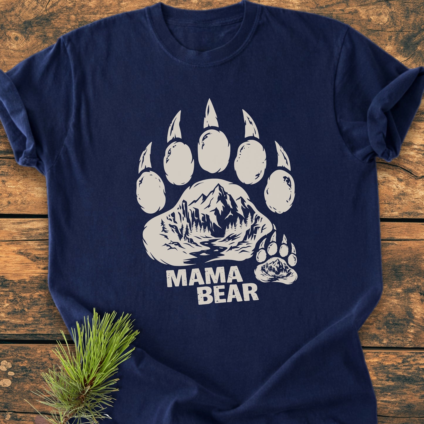 Mama Bear - Vintage Heavyweight Tee featuring a bear's paw print with a mountain landscape and MAMA BEAR text. Laid flat with a pine sprig beside, highlighting its relaxed fit and durability.