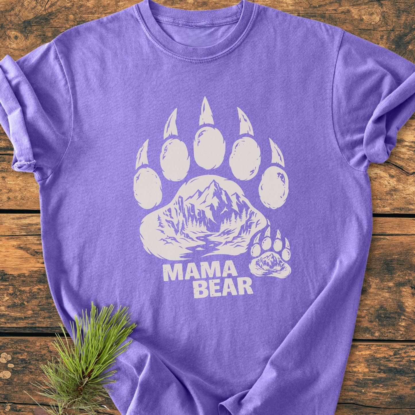 Mama Bear - Vintage Heavyweight Tee features a white bear paw print with a mountain scene on a purple shirt, laid flat on a wooden surface, showcasing its relaxed fit and durable stitching.