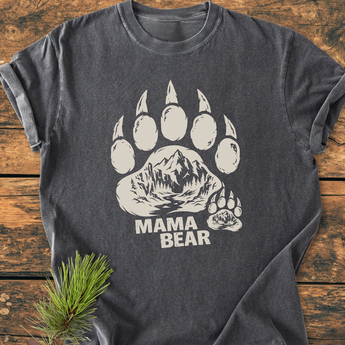 Mama Bear - Vintage Heavyweight Tee features a dark-gray color, short sleeves, a bear-paw print with a mountain-and-river scene, and MAMA BEAR text, laid on a wooden surface with a pine sprig.