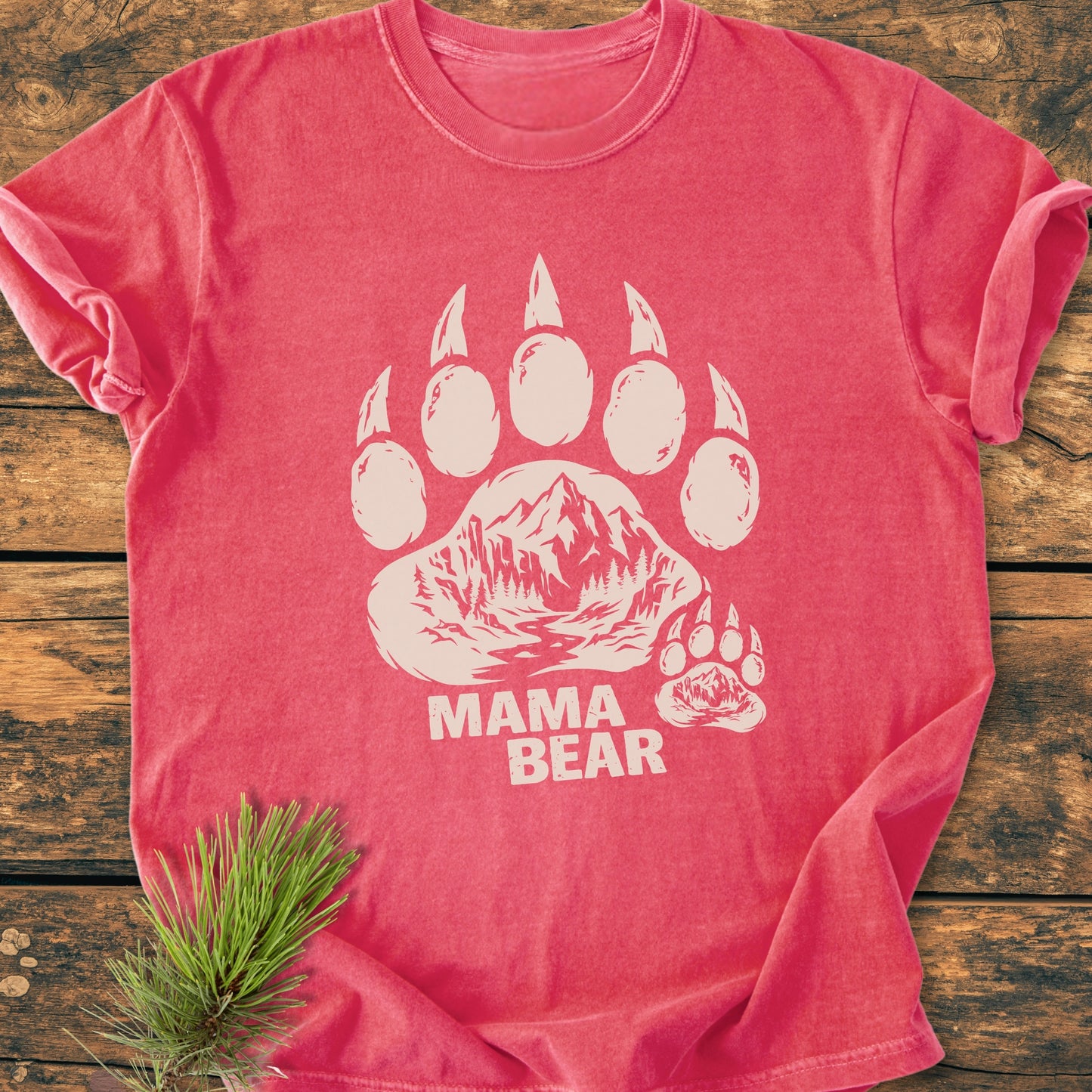 Mama Bear - Vintage Heavyweight Tee, featuring a stylized bear paw print with mountain and river scene, MAMA BEAR text, folded on wood with pine sprig accent.