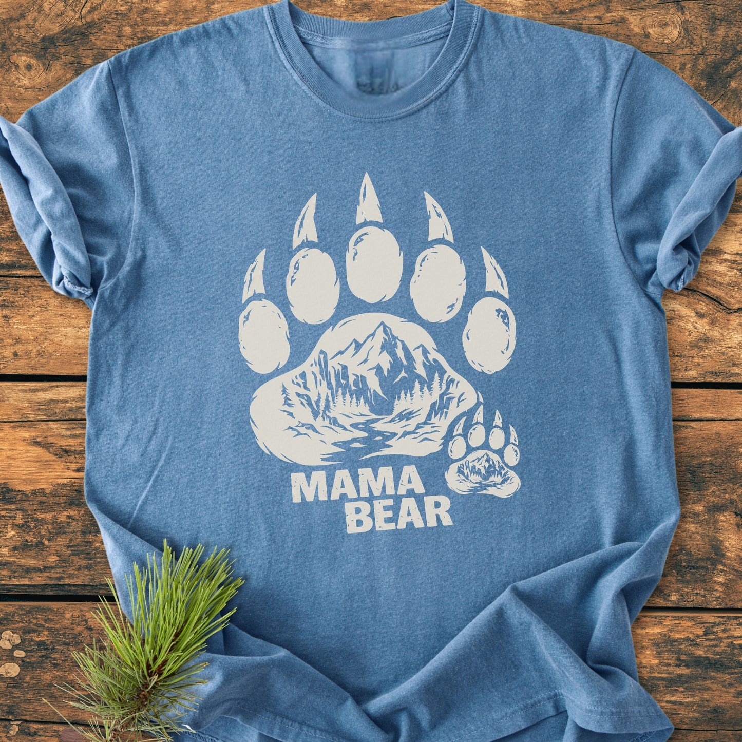 Mama Bear - Vintage Heavyweight Tee featuring a large bear paw with a mountain scene and MAMA BEAR text, laid flat with a pine branch accent, showcasing soft-washed, durable fabric.