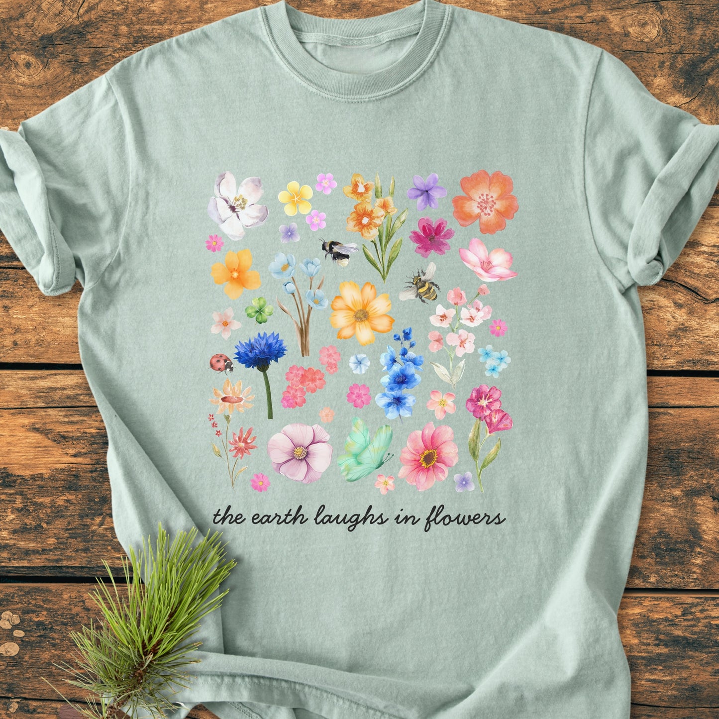 The Earth Laughs In Flowers - Vintage Heavyweight Tee features a watercolor floral grid with insects and script, styled on wood with a pine sprig, showcasing its relaxed fit and cozy fabric.