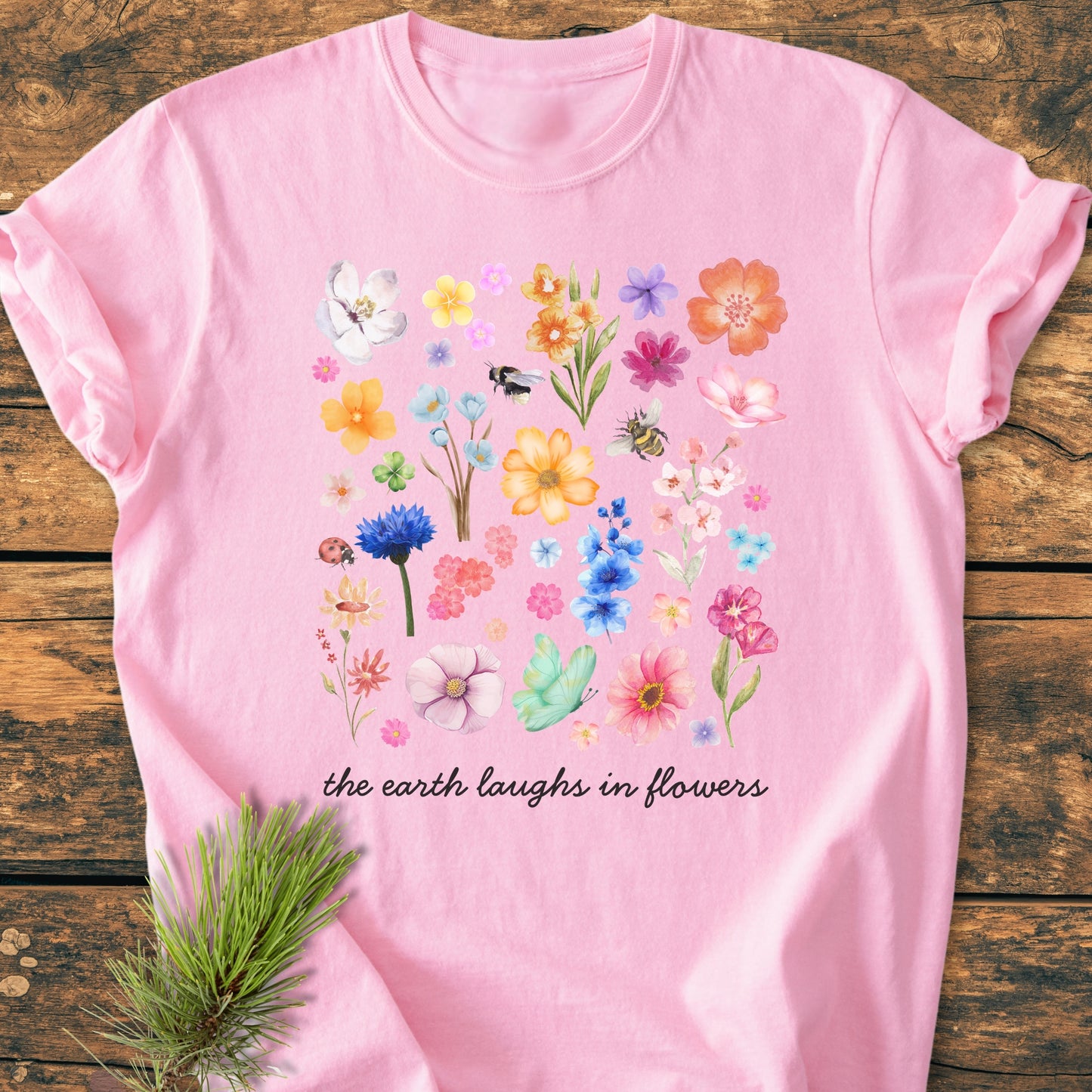 The Earth Laughs In Flowers - Vintage Heavyweight Tee features a watercolor-style floral grid with insects and script text on a soft pink shirt, displayed on rustic wood with a pine sprig.
