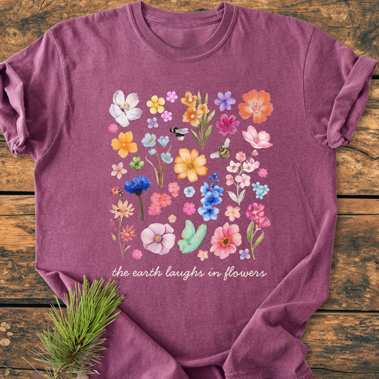 The Earth Laughs In Flowers - Vintage Heavyweight Tee features a colorful floral collage with insects on a heathered-purple crew-neck, styled with a pine sprig on a wooden surface.