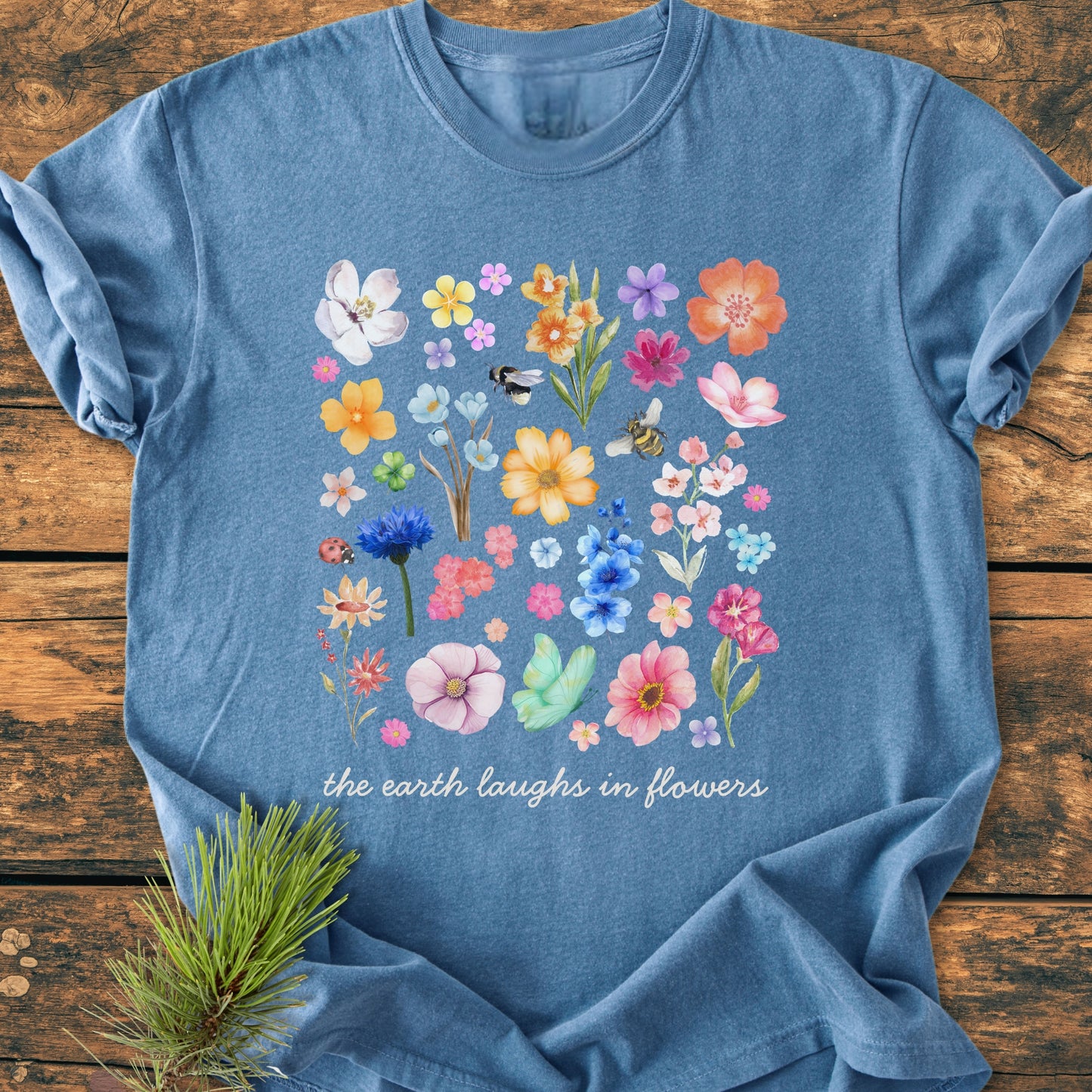 The Earth Laughs In Flowers - Vintage Heavyweight Tee displays a vibrant floral and insect design with a cursive slogan on a soft-blue background, laid out on a wooden surface with a pine sprig beside it.