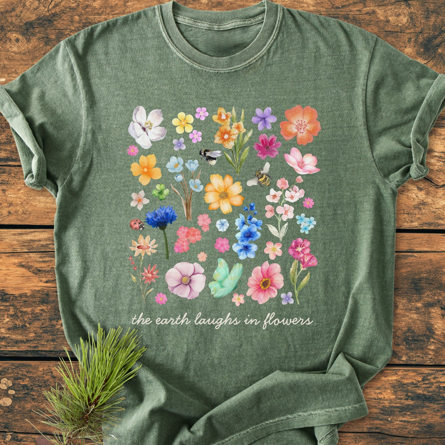 The Earth Laughs In Flowers - Vintage Heavyweight Tee features watercolor-style blooms and insects with the slogan on heather-green fabric, laid on wooden planks with a pine sprig accent.