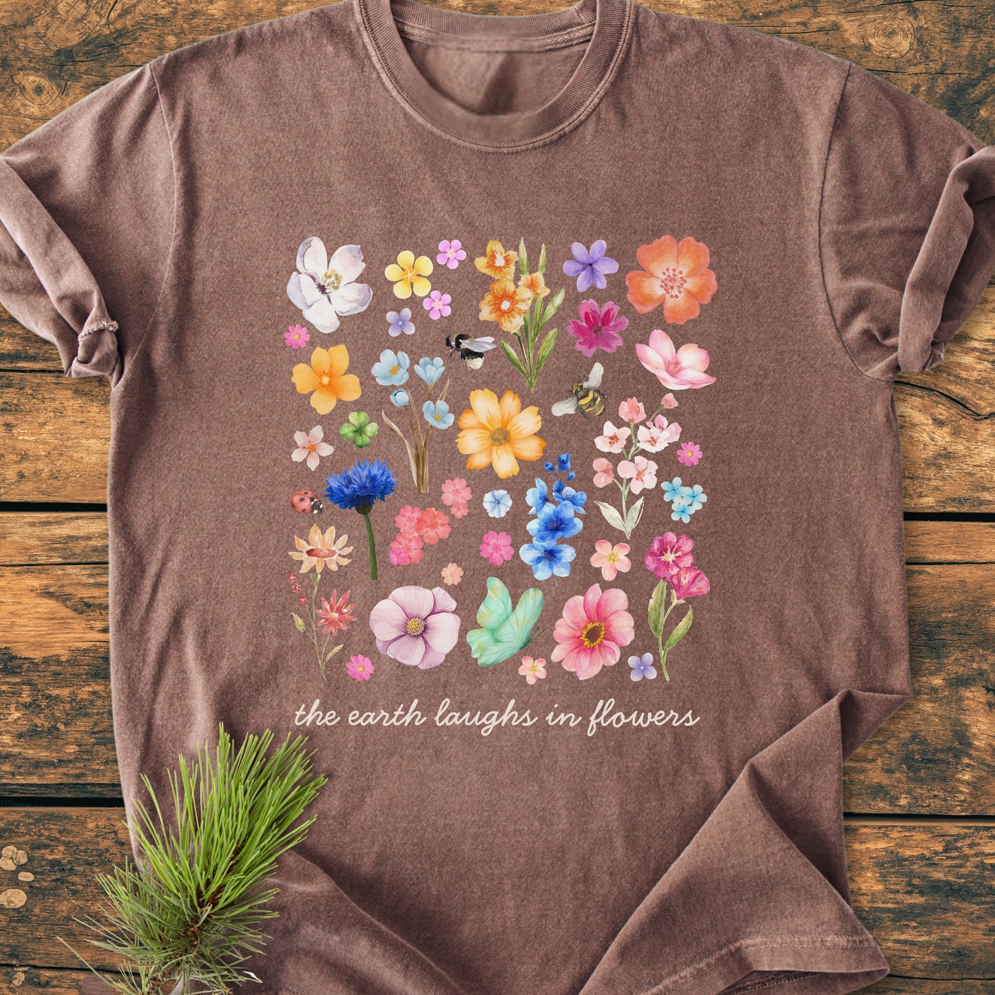 The Earth Laughs In Flowers - Vintage Heavyweight Tee features a hand-painted floral grid with bees and a ladybug, and a cursive quote, on a rustic wooden surface.