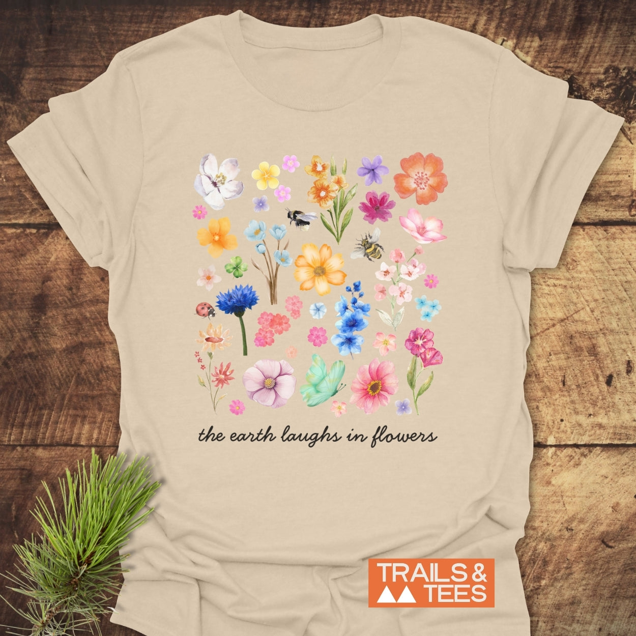 The Earth Laughs In Flowers T-Shirt features a grid of watercolor flowers, bees, and a ladybug with the slogan below. Crafted from soft cotton, styled with pine needles, and branded “TRAILS & TEES.”
