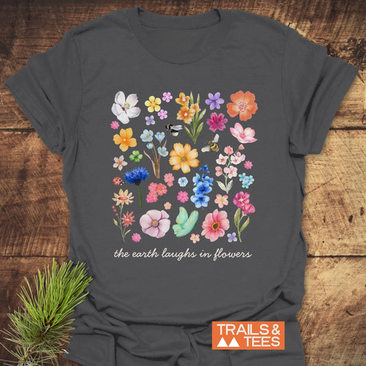 The Earth Laughs In Flowers T-Shirt displays vibrant floral illustrations with bees and a ladybug on dark gray cotton, featuring the phrase in cursive, styled with a pine sprig and Trails & Tees logo.