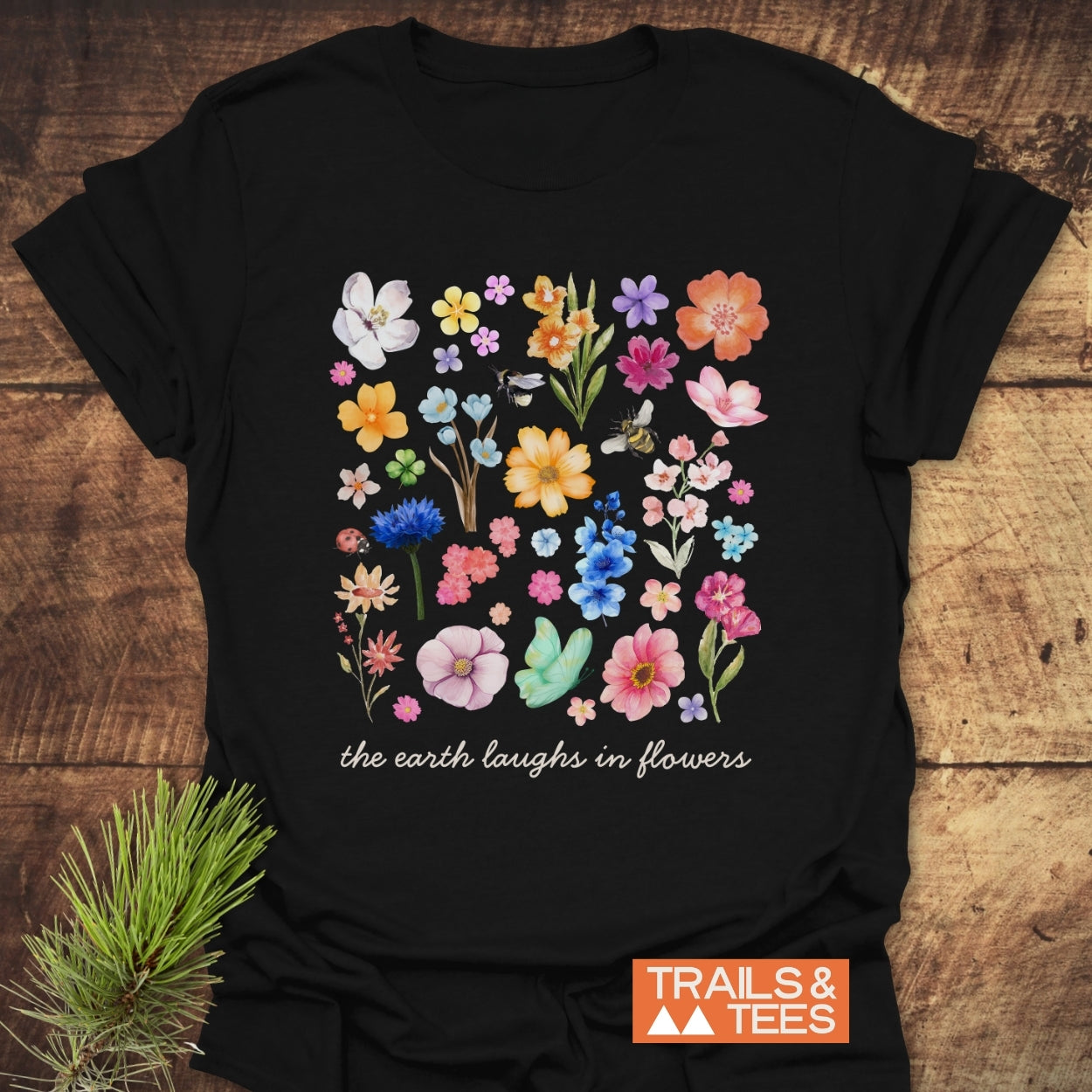 The Earth Laughs In Flowers T-Shirt features a colorful watercolor-style floral collage with insects and the slogan beneath. Showcases a Trails & Tees logo, ensuring trail-tested comfort and durability.