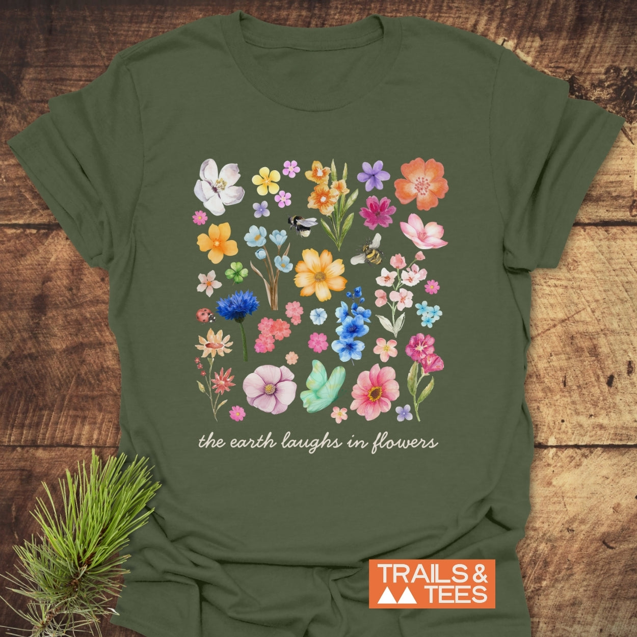 The Earth Laughs In Flowers T-Shirt on rustic wood, showcasing a floral grid with bees and a ladybug; features “the earth laughs in flowers” slogan. Includes TRAILS & TEES logo and pine sprig.