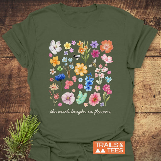 The Earth Laughs In Flowers T-Shirt on rustic wood, showcasing a floral grid with bees and a ladybug; features “the earth laughs in flowers” slogan. Includes TRAILS & TEES logo and pine sprig.