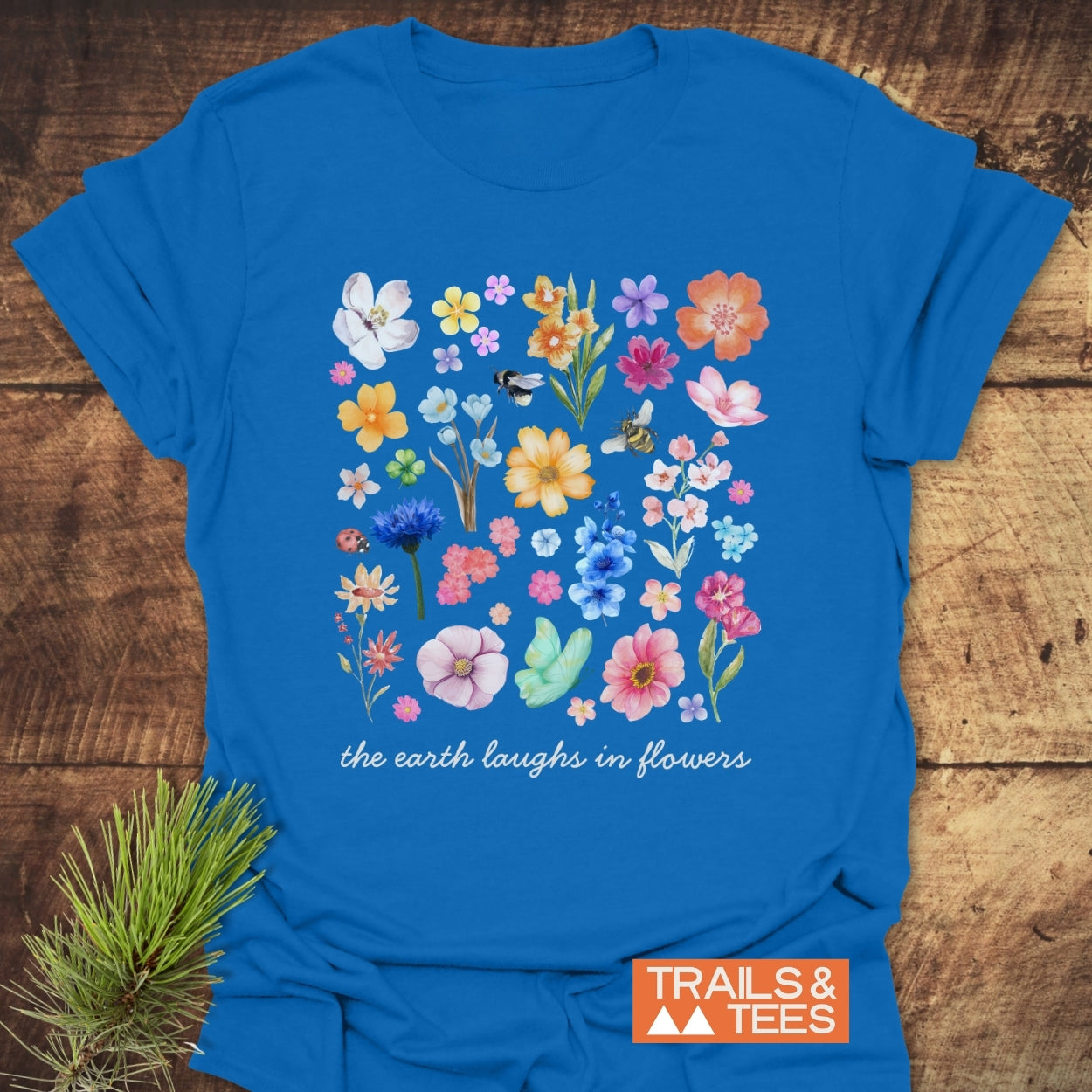 The Earth Laughs In Flowers T-Shirt features a grid of watercolor-style flowers, a bee, and a ladybug, with the phrase beneath. An orange TRAILS & TEES logo is visible in the corner.
