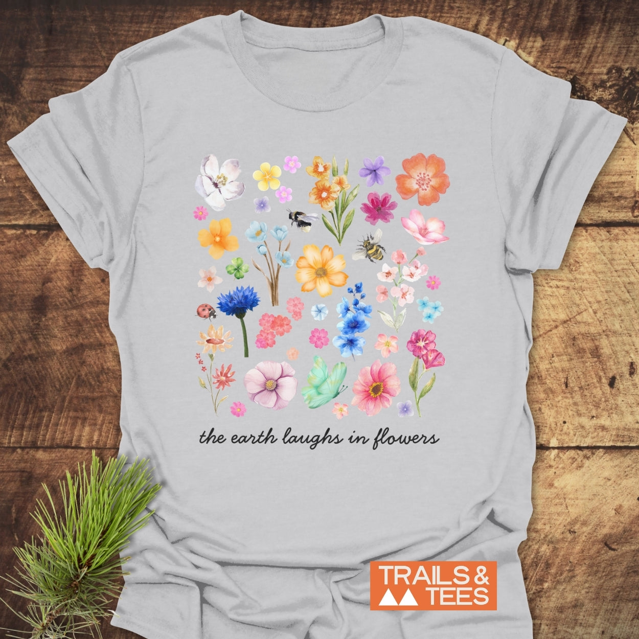 The Earth Laughs In Flowers T-Shirt features a floral watercolor grid with bees and a ladybug, accented by a cursive quote. Laid flat, it showcases Trails & Tees branding, embodying trail-tested comfort.