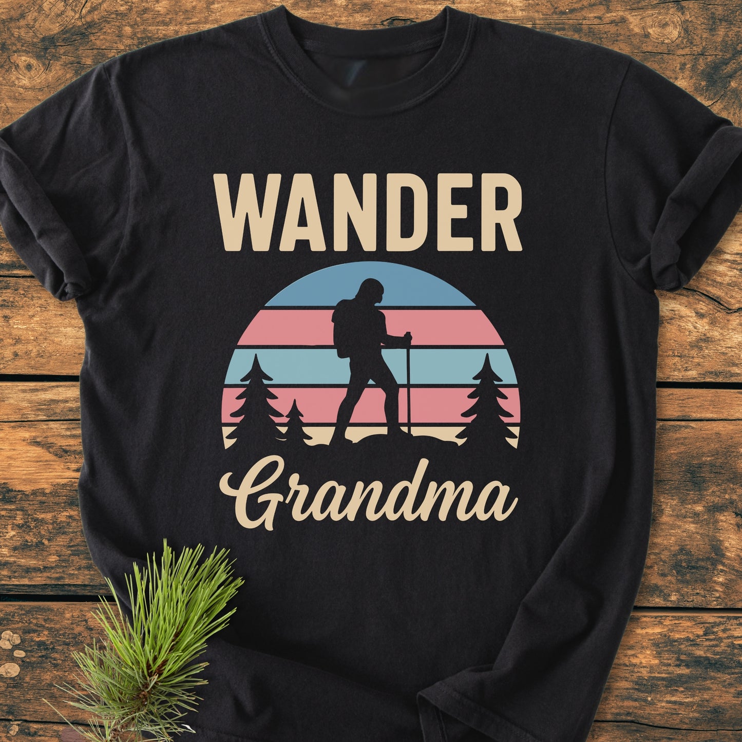 Wander Grandma - Vintage Heavyweight Tee features a retro sunset graphic with a backpacker silhouette and pine trees. Laid on wood with a pine sprig, highlighting its cozy, relaxed fit.
