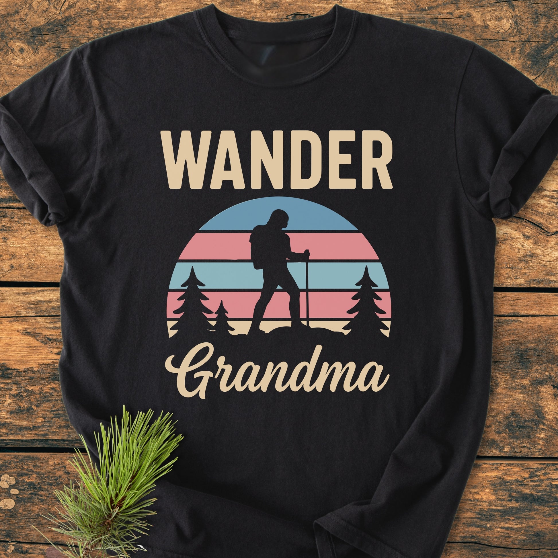 Wander Grandma - Vintage Heavyweight Tee features a retro sunset graphic with a backpacker silhouette and pine trees. Laid on wood with a pine sprig, highlighting its cozy, relaxed fit.