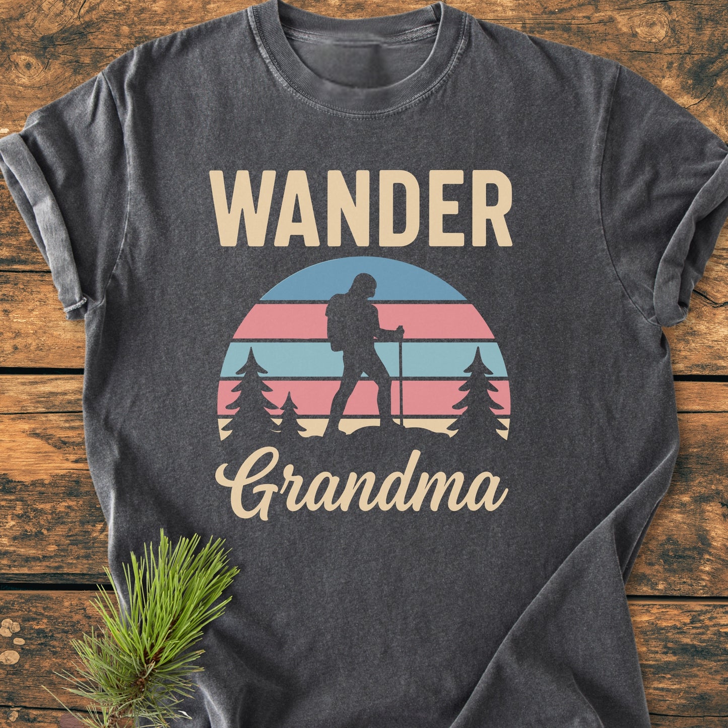 Wander Grandma - Vintage Heavyweight Tee: Charcoal-gray T-shirt with retro graphic of a hiking scene and WANDER Grandma text, laid on rustic wood with pine sprig. Cozy, durable, 100% ring-spun cotton.