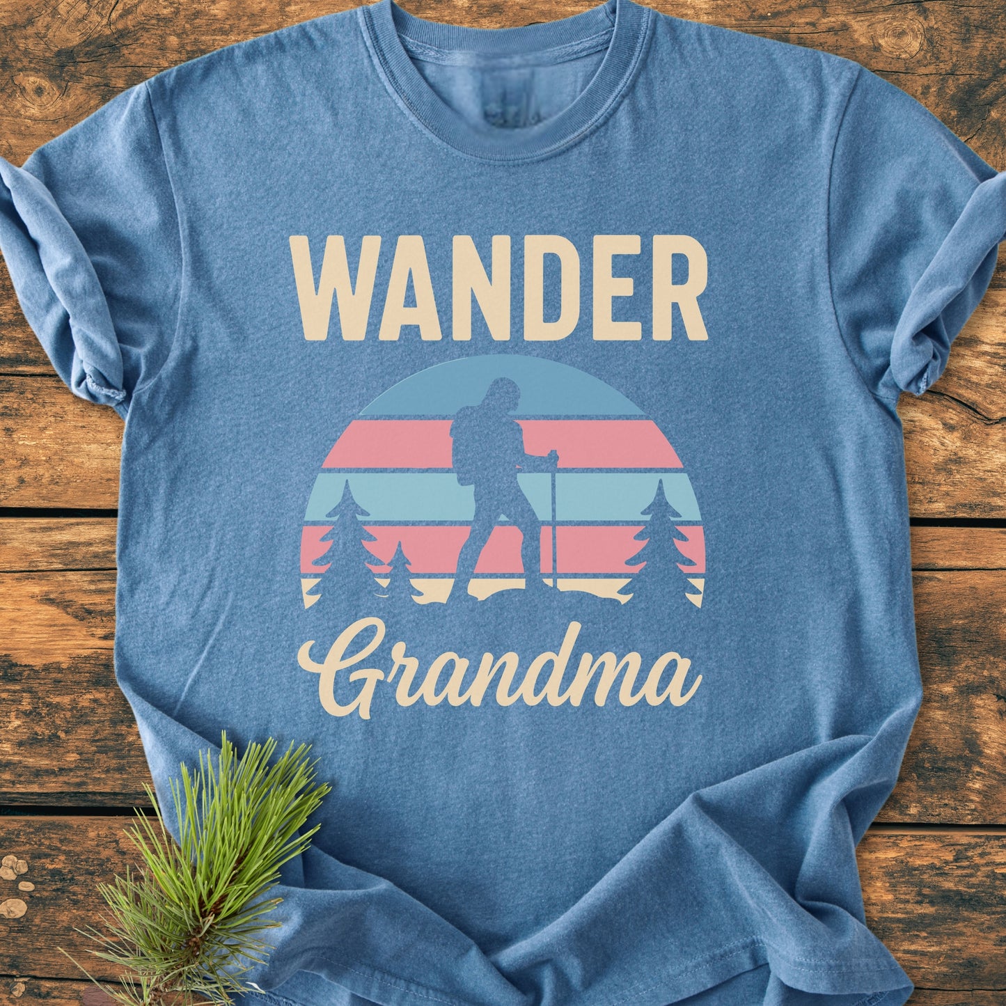 Heather-blue Wander Grandma - Vintage Heavyweight Tee featuring a hiker silhouette against a striped sunset, with bold WANDER and script Grandma text. Laid on wood with pine sprig accent.