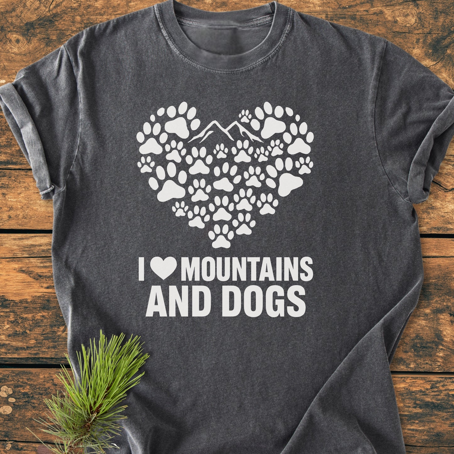 The Paw Heart Dog Lover - Vintage Heavyweight Tee features a heart of paw prints and mountain silhouette, with “I ♥ MOUNTAINS AND DOGS” text on a charcoal-gray T-shirt, displayed on a wooden surface.