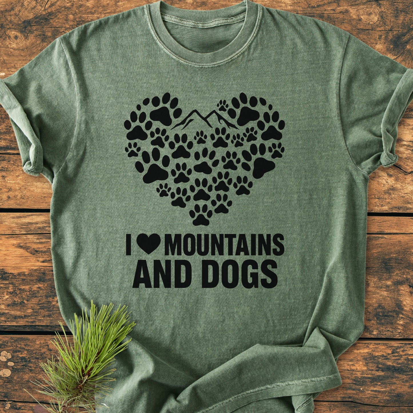 Paw Heart Dog Lover - Vintage Heavyweight Tee features a heart-shaped dog paw print graphic with a mountain peak, on a heather-green T-shirt, laid on a wooden surface with a pine sprig.