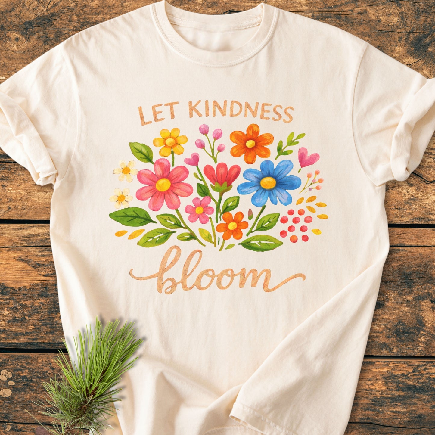The Let Kindness Bloom - Vintage Wash Tee features a watercolor floral design and uplifting text on a cream T-shirt, shown on a rustic wooden background with a pine sprig nearby.