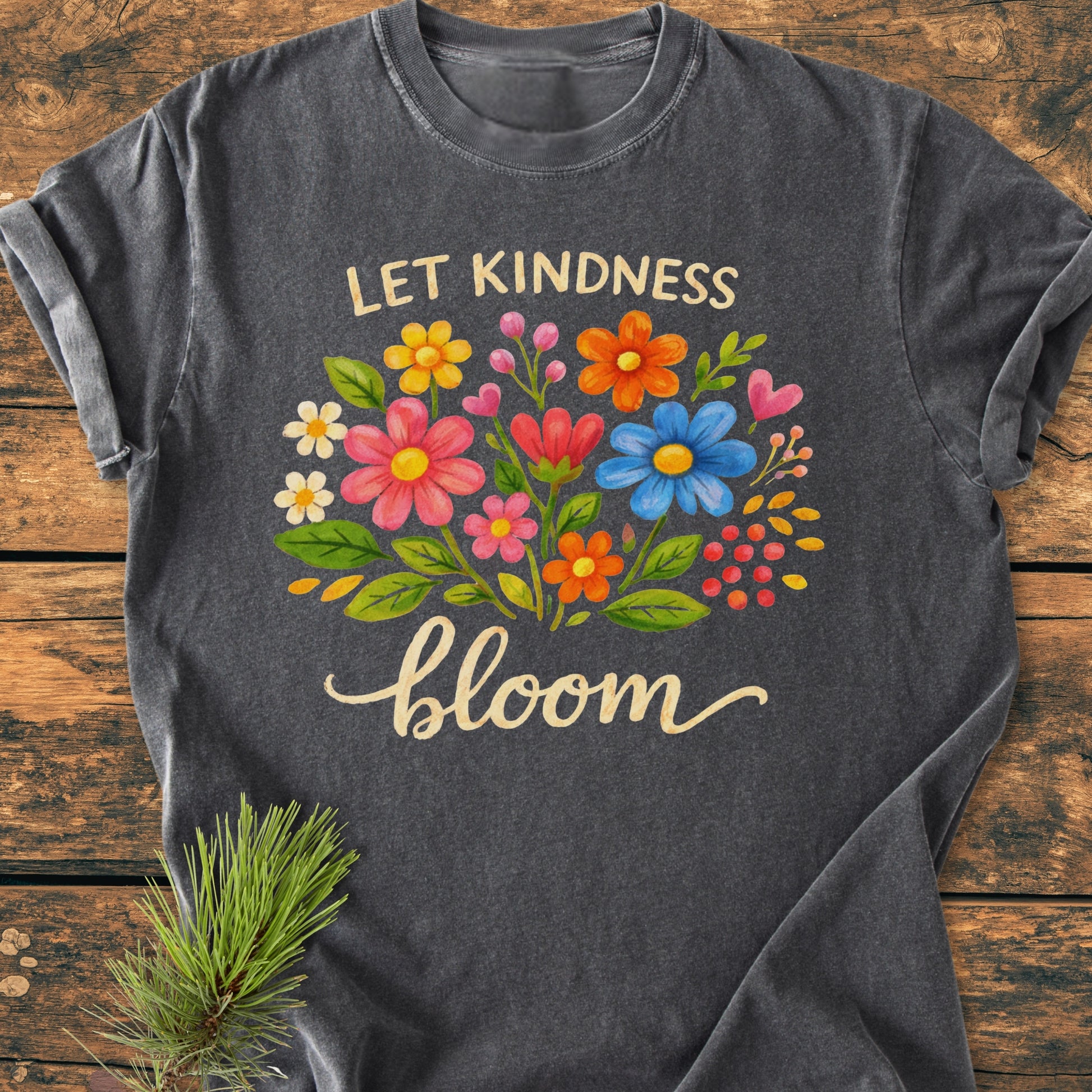 Let Kindness Bloom - Vintage Wash Tee featuring a vibrant floral design and script, displayed on a rustic wooden surface with a pine sprig beside, highlighting its cozy, relaxed fit and durability.