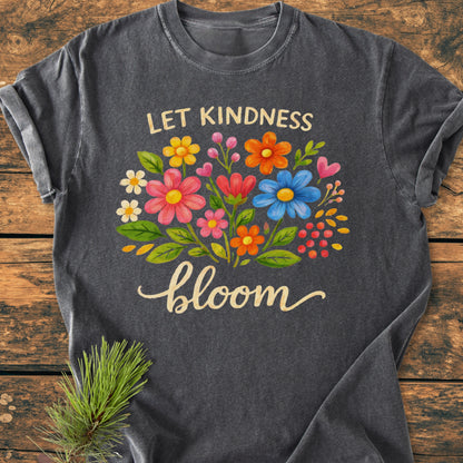 Let Kindness Bloom - Vintage Wash Tee featuring a vibrant floral design and script, displayed on a rustic wooden surface with a pine sprig beside, highlighting its cozy, relaxed fit and durability.