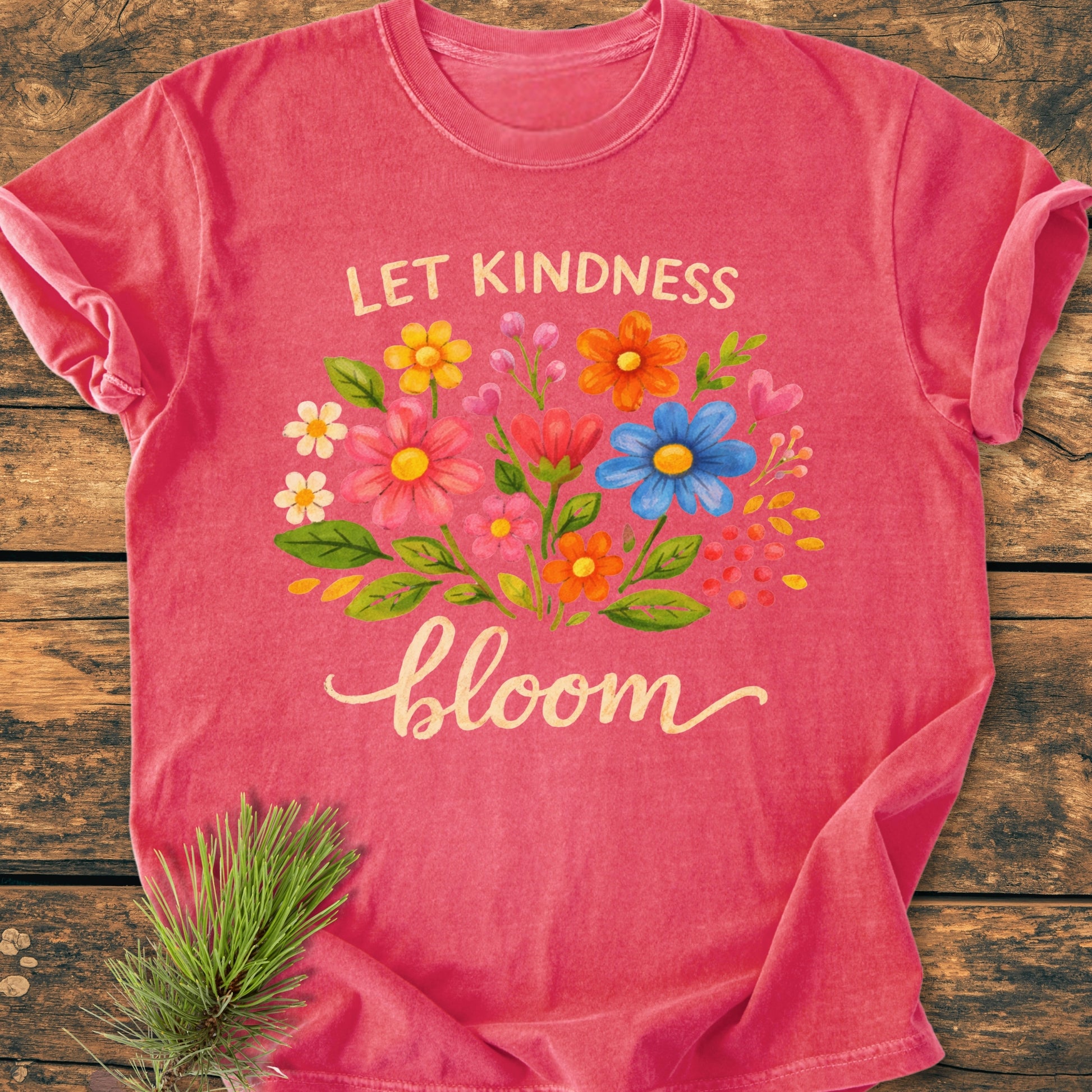 Let Kindness Bloom - Vintage Wash Tee featuring a colorful floral graphic and LET KINDNESS bloom text on coral-pink fabric, laid flat on rustic wood beside a pine sprig.