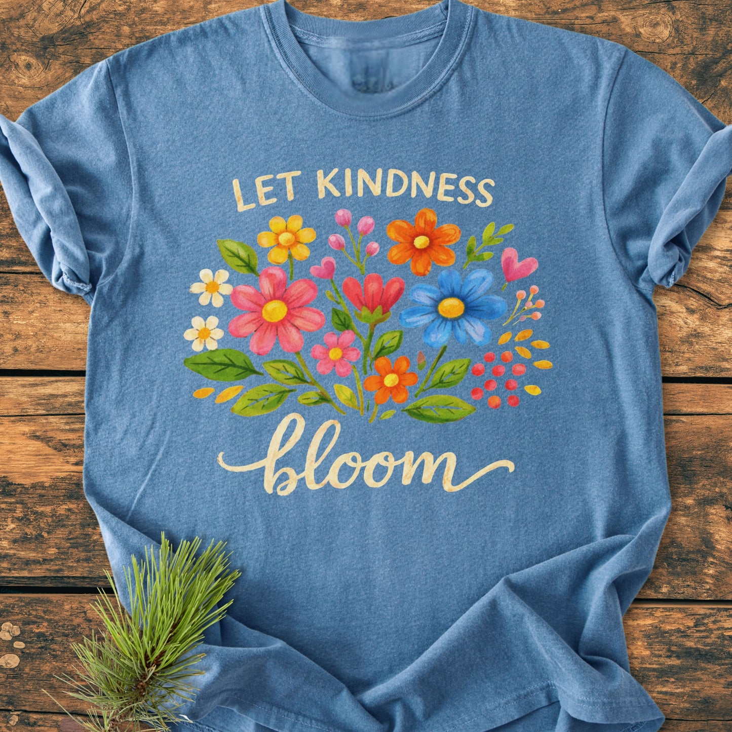 Let Kindness Bloom - Vintage Wash Tee, heathered-blue, features a vibrant floral design with “LET KINDNESS” and “bloom” text, laid on wood. Enhanced with pine needles, showcasing its cozy and relaxed style.