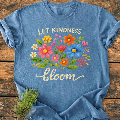 Let Kindness Bloom - Vintage Wash Tee, heathered-blue, features a vibrant floral design with “LET KINDNESS” and “bloom” text, laid on wood. Enhanced with pine needles, showcasing its cozy and relaxed style.