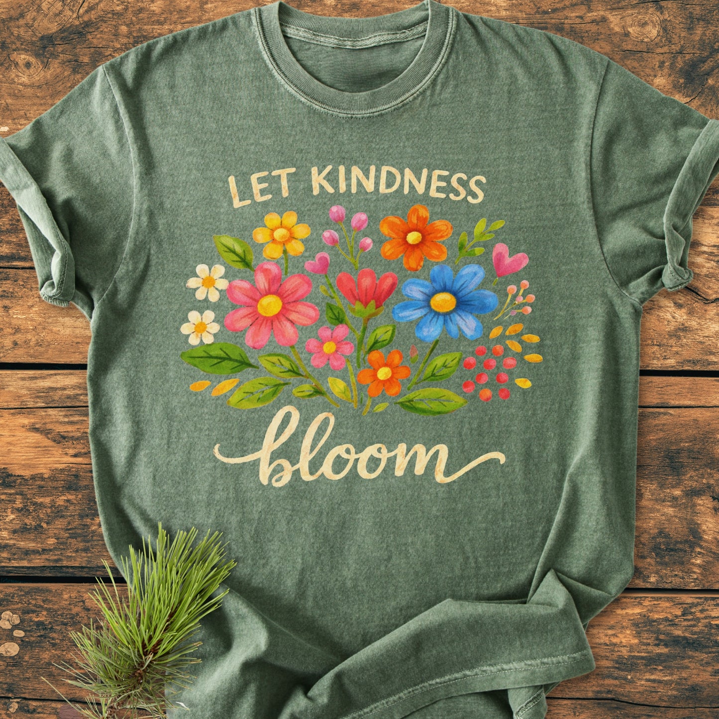 The Let Kindness Bloom - Vintage Wash Tee features a floral design with uplifting text on an earthy green background, laid flat on a wooden surface, exemplifying comfort and style.
