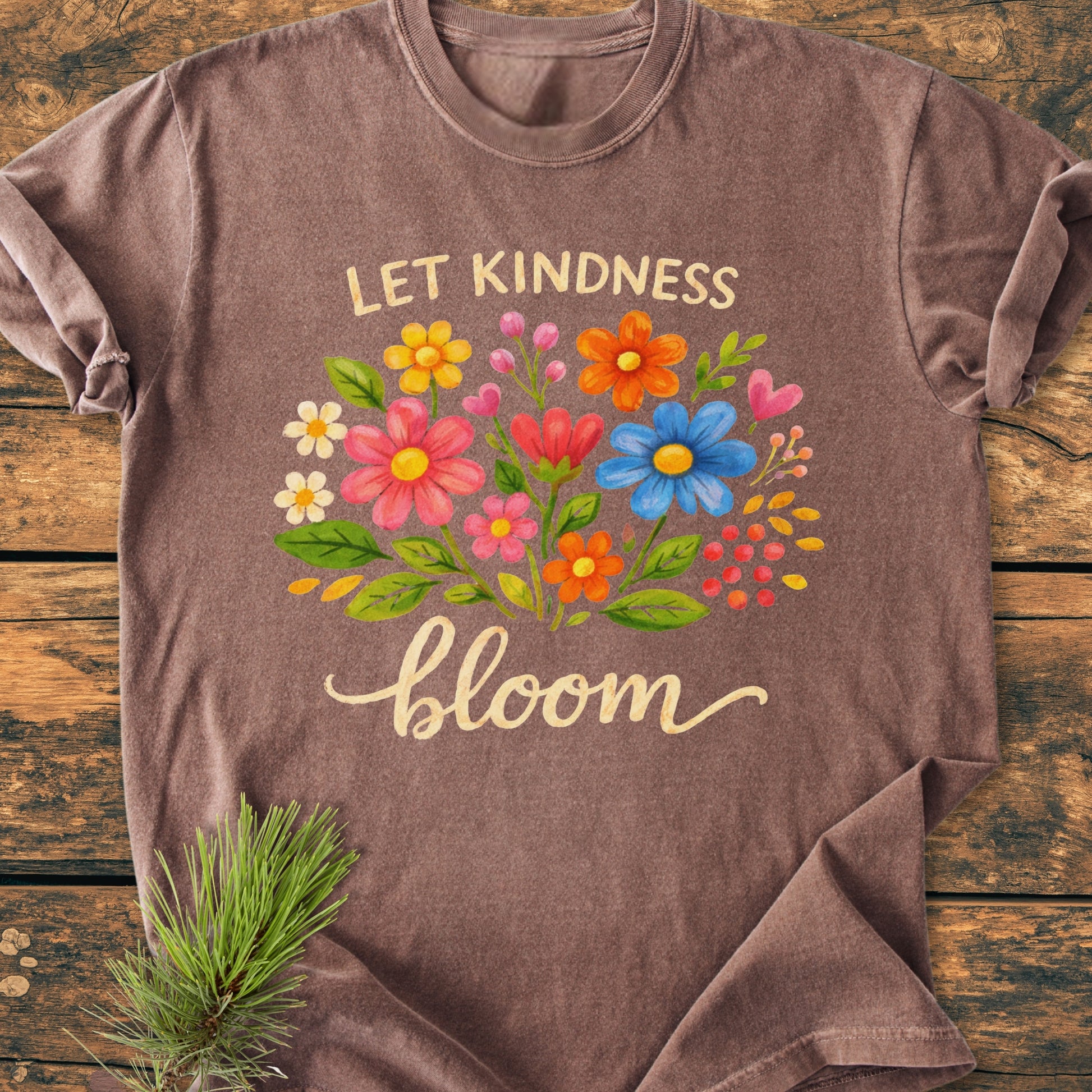 Let Kindness Bloom - Vintage Wash Tee on wooden surface, features a vibrant floral design and the phrase “LET KINDNESS bloom” in artistic fonts, showcasing its relaxed fit and durable fabric.