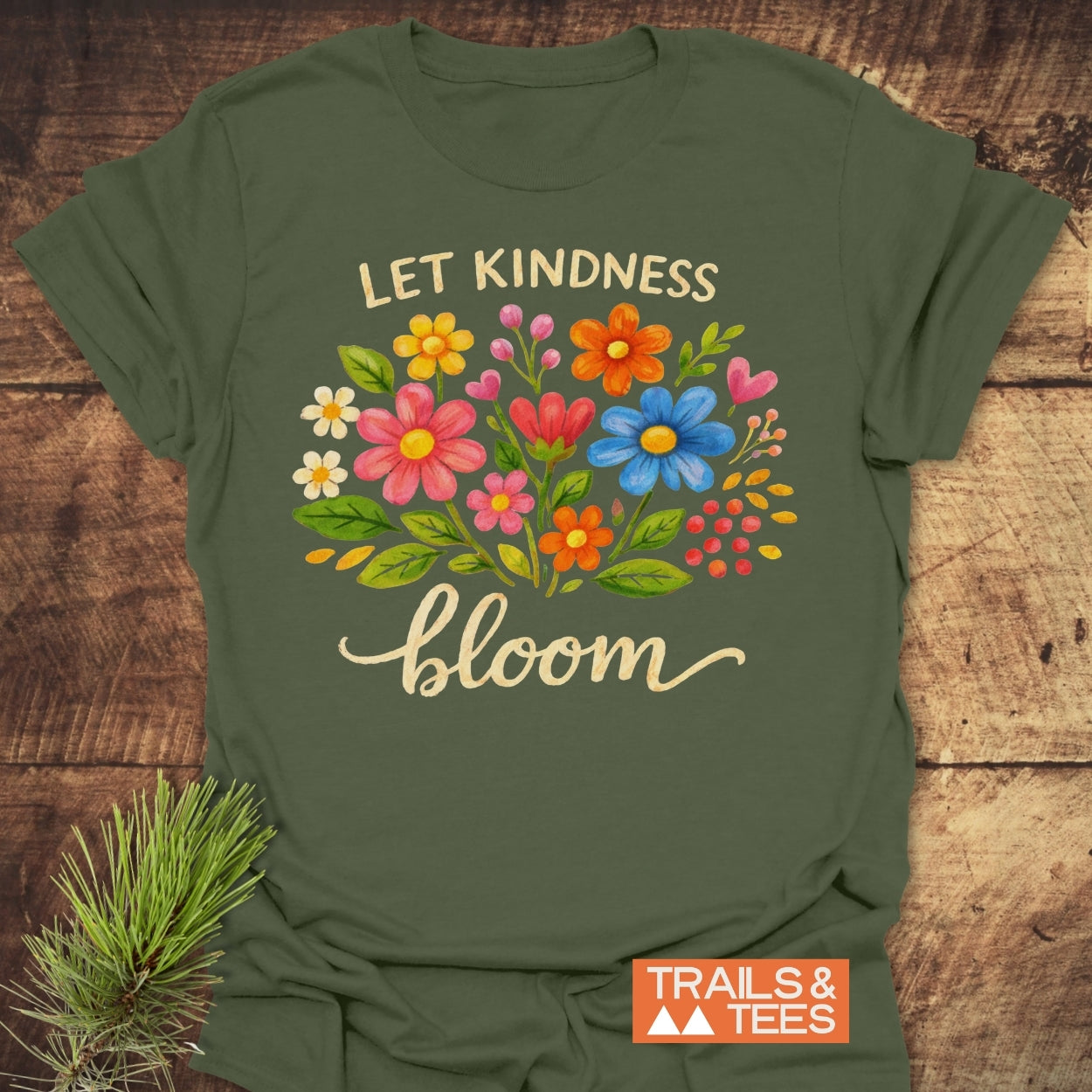 Let Kindness Bloom T-Shirt featuring a vibrant floral design and inspiring text, on soft ring-spun cotton, showcasing outdoor spirit with a Trails & Tees logo, laid on a wooden surface.
