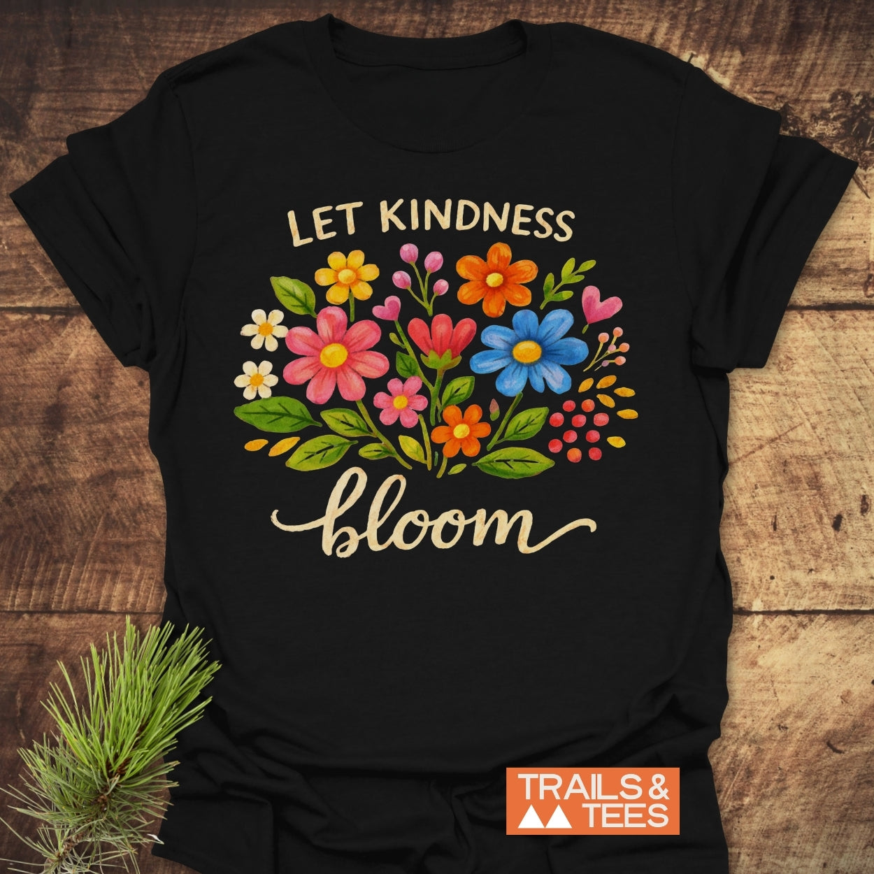 Let Kindness Bloom T-Shirt with vibrant floral print and motivational text, crafted from soft cotton, ideal for outdoor enthusiasts. Features durable design with pre-shrunk fit and Trails & Tees logo.