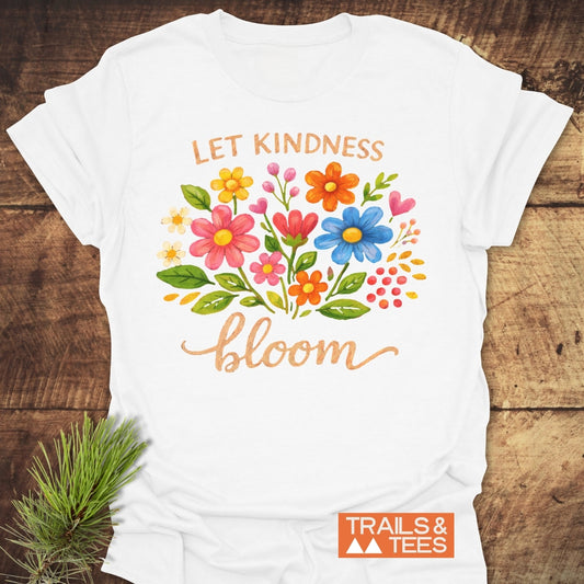 Let Kindness Bloom T-Shirt with watercolor flowers and 'LET KINDNESS bloom' text. Soft ring-spun cotton, orange Trails & Tees logo, styled with pine sprig on a wooden surface.