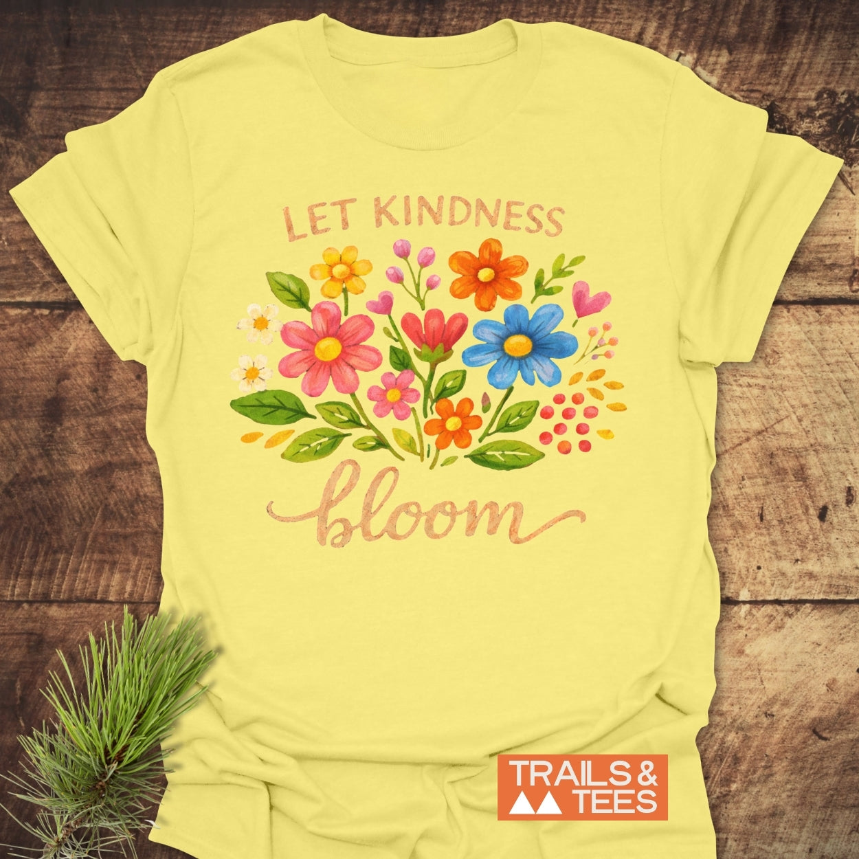 Let Kindness Bloom T-Shirt featuring a hand-painted floral bouquet with the phrase “LET KINDNESS bloom” in warm font, displayed on a rustic surface with a pine sprig beside it.