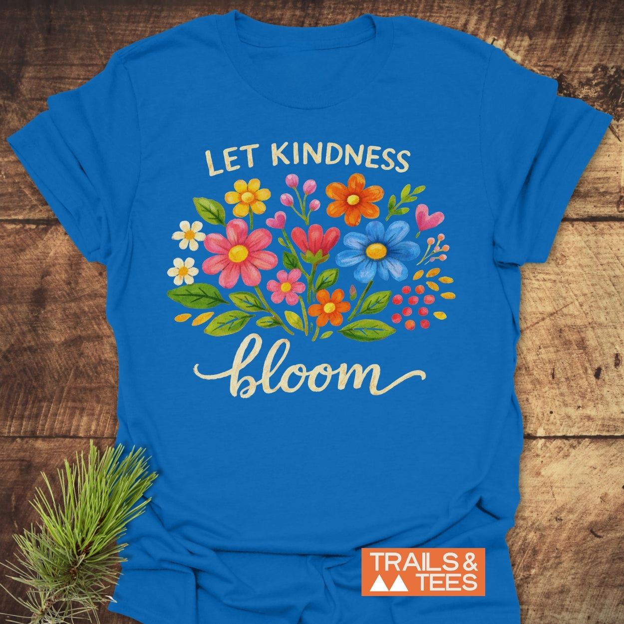 The Let Kindness Bloom T-Shirt features a floral design with uplifting text, displayed on a wooden surface beside a pine sprig, capturing its rustic charm and outdoor spirit.